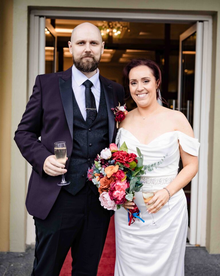 Bride and groom smiling at the camera, holding champagne and a bright bouquet at the door of Vienna Woods Hotel, Cork