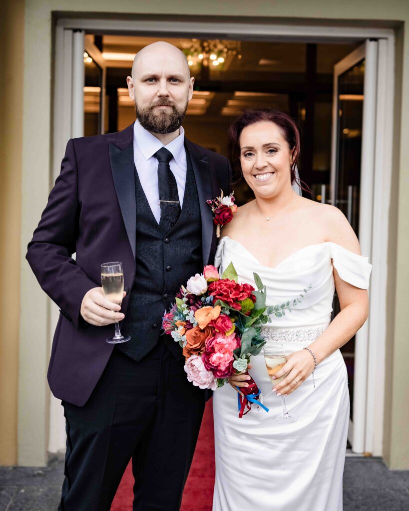 Bride and groom holding champagne glasses and a bright bouquet at the door of the reception venue, groom in a purple dinner jacket