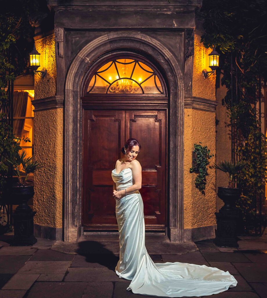 Sinéad in a satin gown with a long train standing in front of an arched wooden door at night, Vienna Woods Hotel wedding, Cork