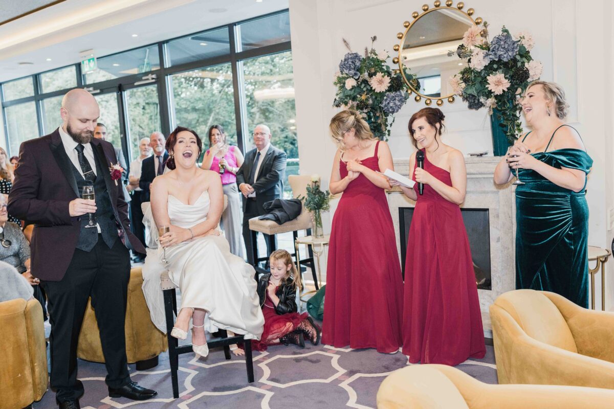 Groom standing with a drink during the speeches while the bride laughs beside bridesmaids in red at Vienna Woods Hotel, Cork