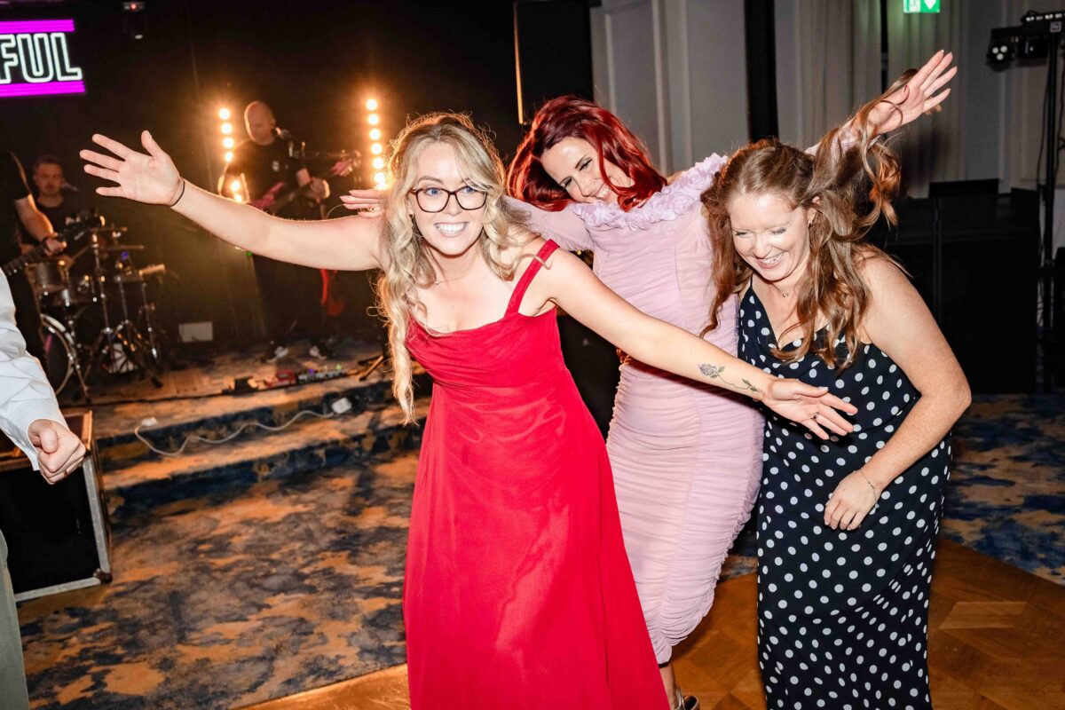 Three guests in red, pink and polka dot dresses dancing with arms out in front of the band The Wilful at a Cork wedding