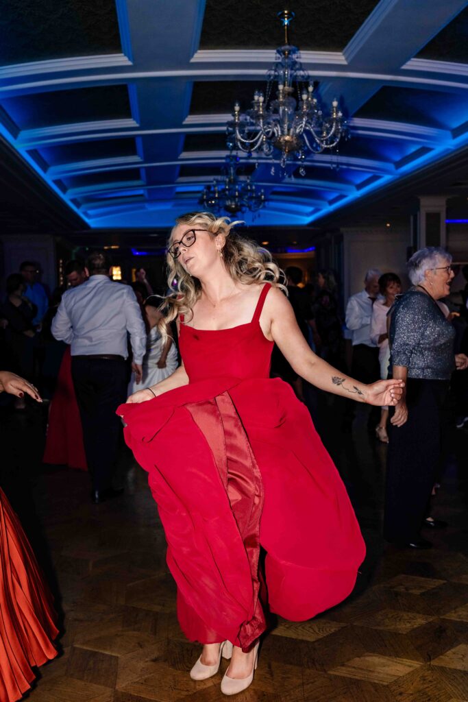 Wedding guest in a red dress with glasses swirling her skirt on the dance floor under blue chandelier lighting