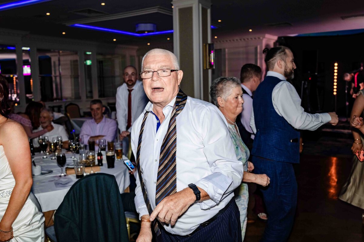 Older guest with his tie loosened dancing and laughing at the wedding reception while others watch from the tables