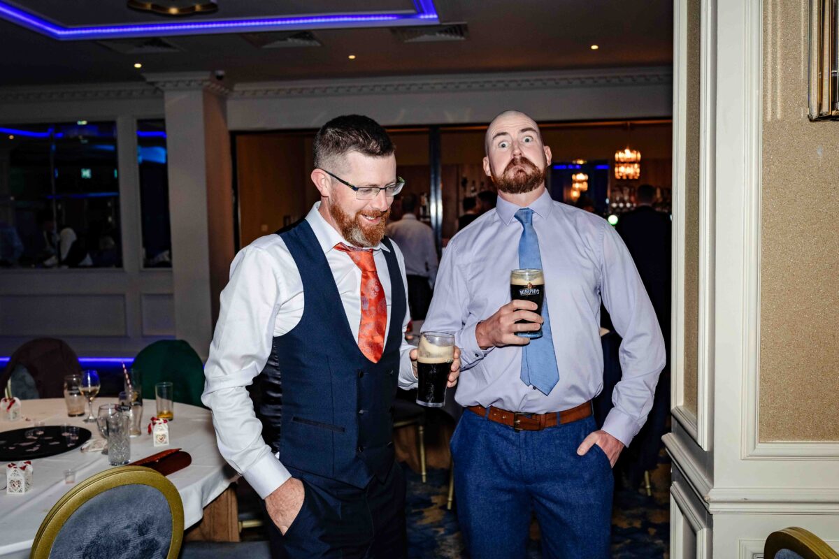 Two groomsmen chatting at the wedding reception, one holding a pint of stout, beside a tall white pillar