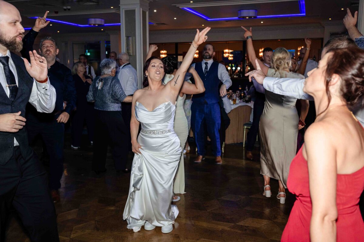 Bride in a satin wedding dress with her arm raised high dancing with guests at Vienna Woods Hotel, Cork