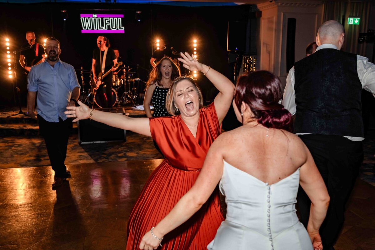 Guests dancing in front of the Wilful wedding band on stage, with a woman in an orange dress and the bride in white