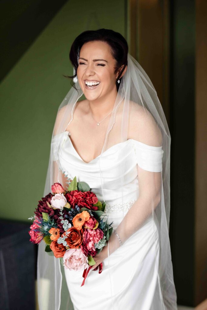 Bride laughing with her head tilted, holding a colourful autumn-toned bouquet beside a green wall