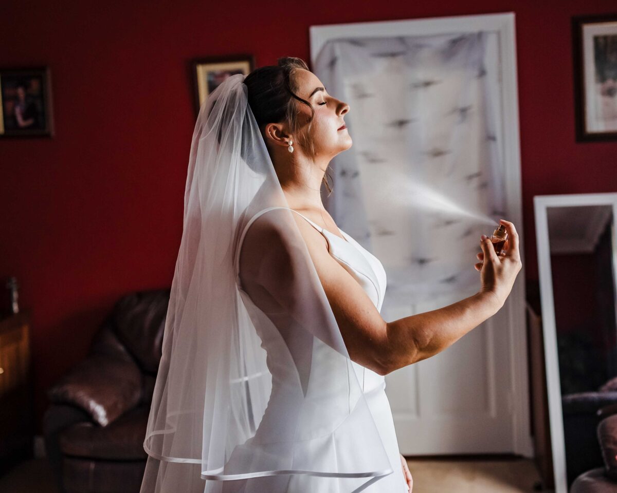Bride in a long veil and white dress tilting her head back as she is spritzed with perfume against a deep red wall on the wedding morning