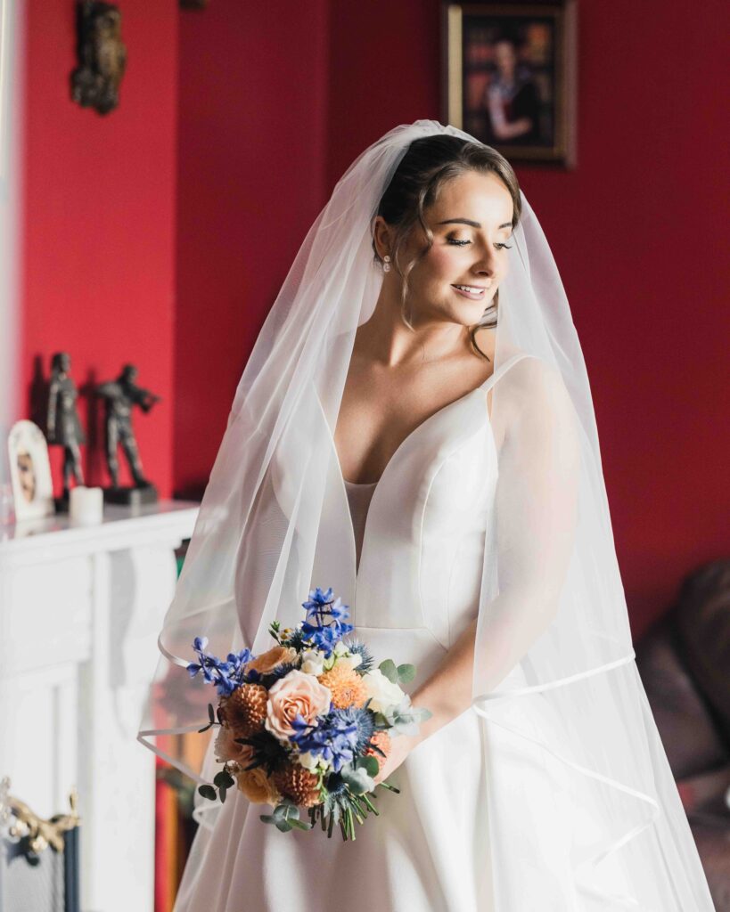 Bride in a veil and V-neck wedding dress holding an orange and blue bouquet beside a mantelpiece in a red room