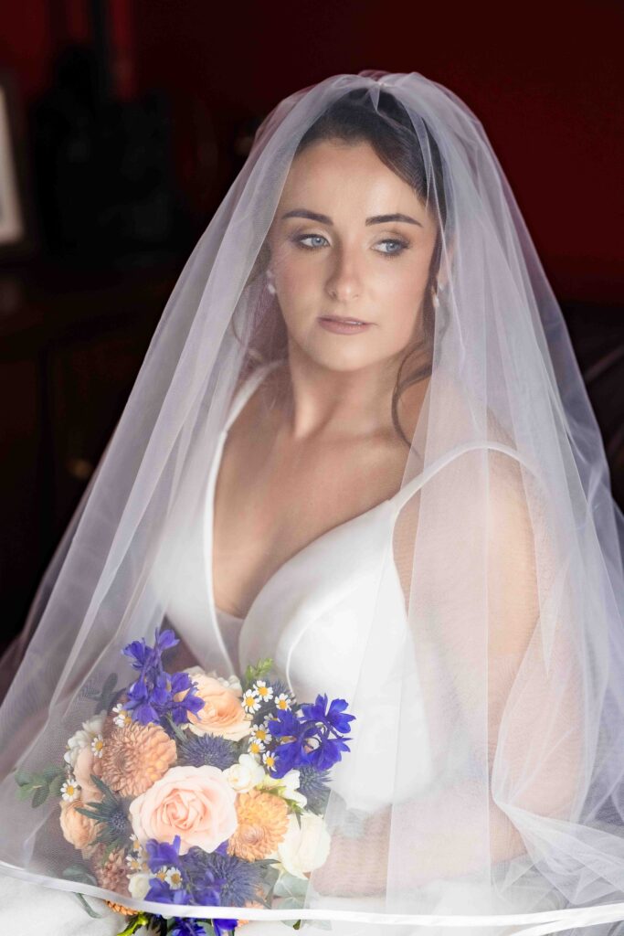 Bride looking off to the side through a sheer veil, holding a peach and blue bouquet in a red-walled room before the ceremony
