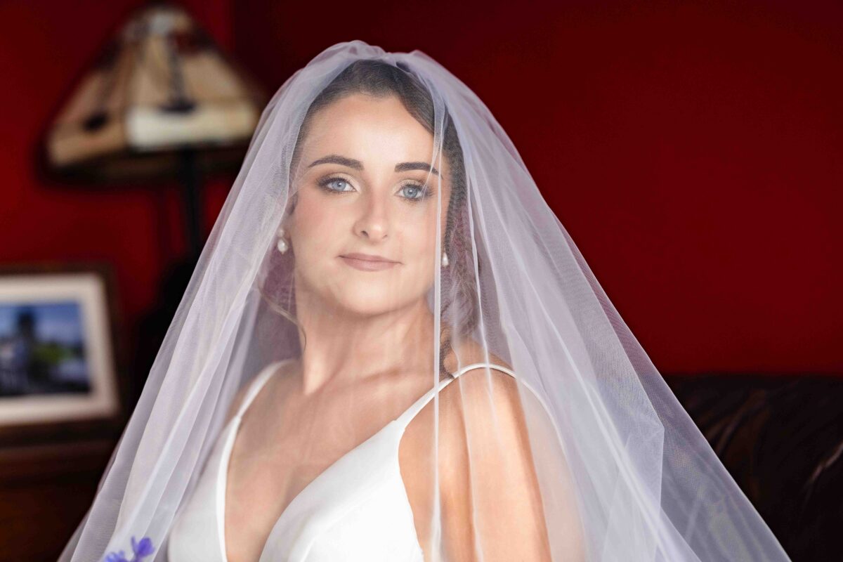 Bride in a satin dress looking straight at the camera through her veil in front of a red wall on the wedding morning