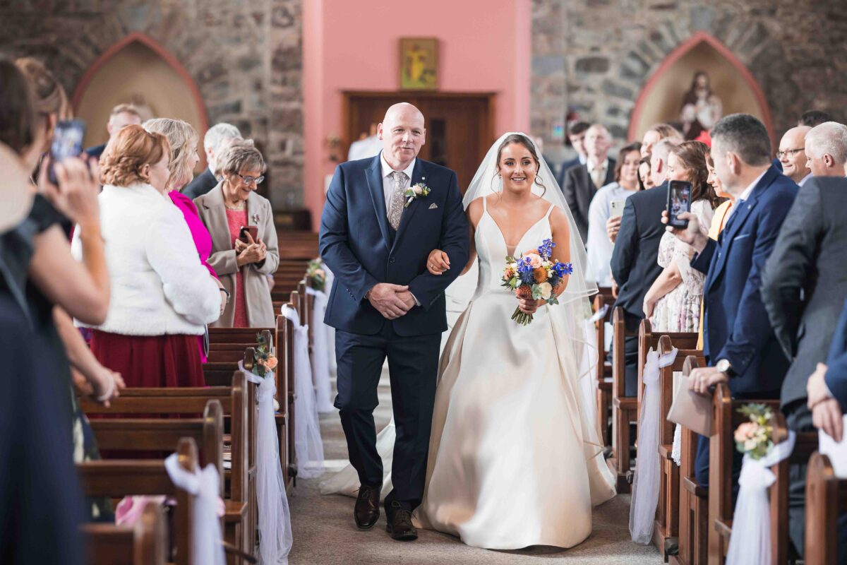 Bride walking up the aisle on her father's arm past guests and wooden pews in Derrinagree Church, Co. Cork