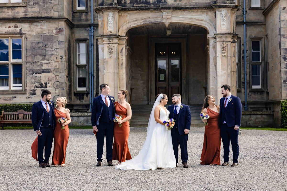 Bride and groom with bridesmaids in rust dresses and groomsmen in navy suits standing at the entrance of Muckross House, Killarney