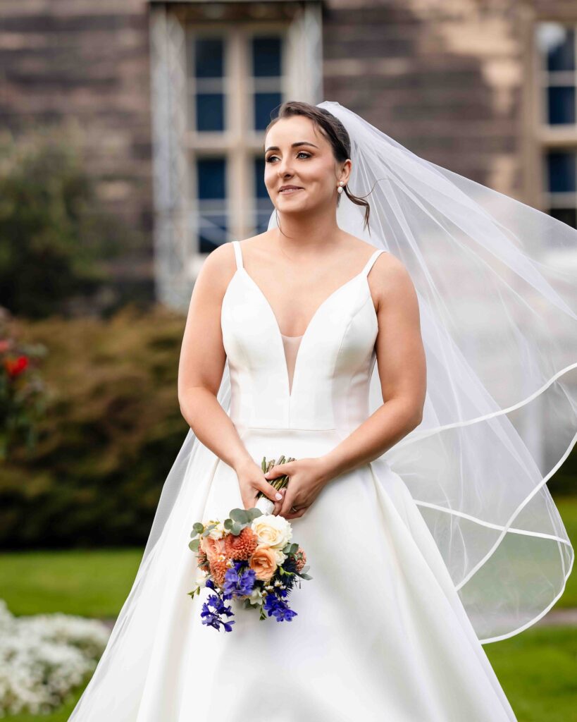 Smiling bride with a peach and blue bouquet and a flowing veil in front of the stone walls of Muckross House, looking off to the side