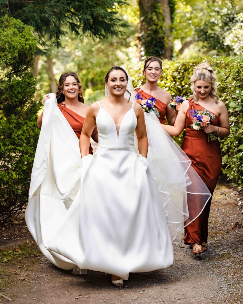 Bride walking through the gardens at Muckross House with three bridesmaids in rust dresses helping carry her long train