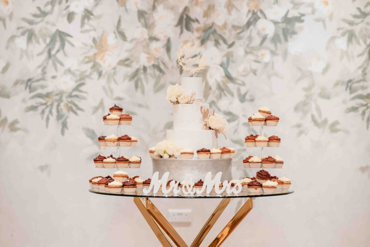 White wedding cake and rust-topped cupcakes on a gold stand with a Mr and Mrs sign against a floral backdrop