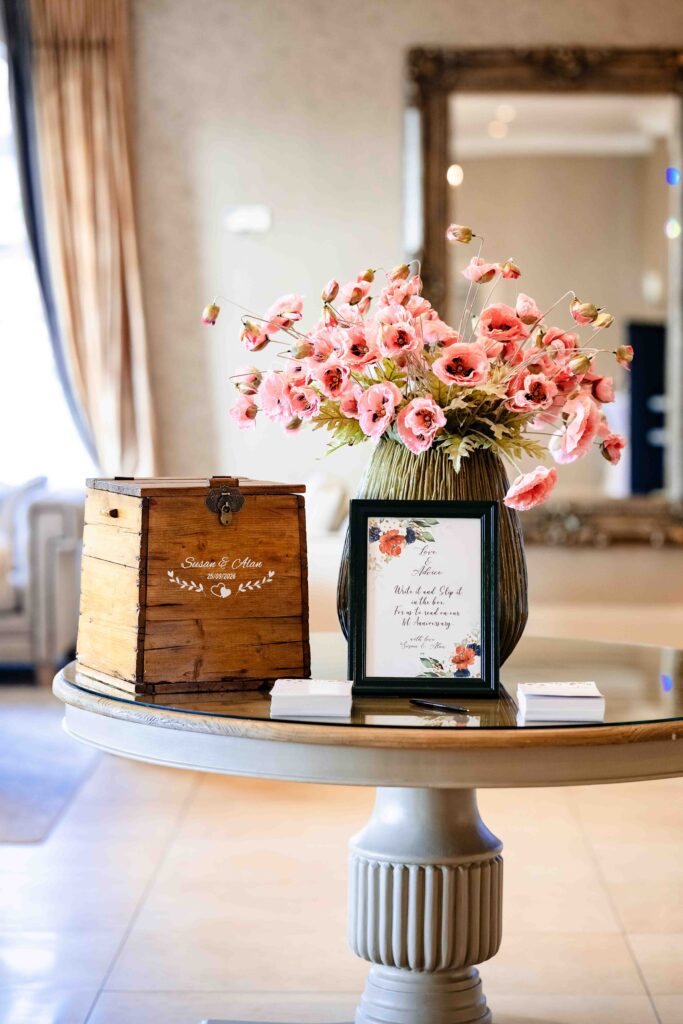 Wedding card box and framed sign on a round table beside a vase of pink flowers at The Oaks, Killarney