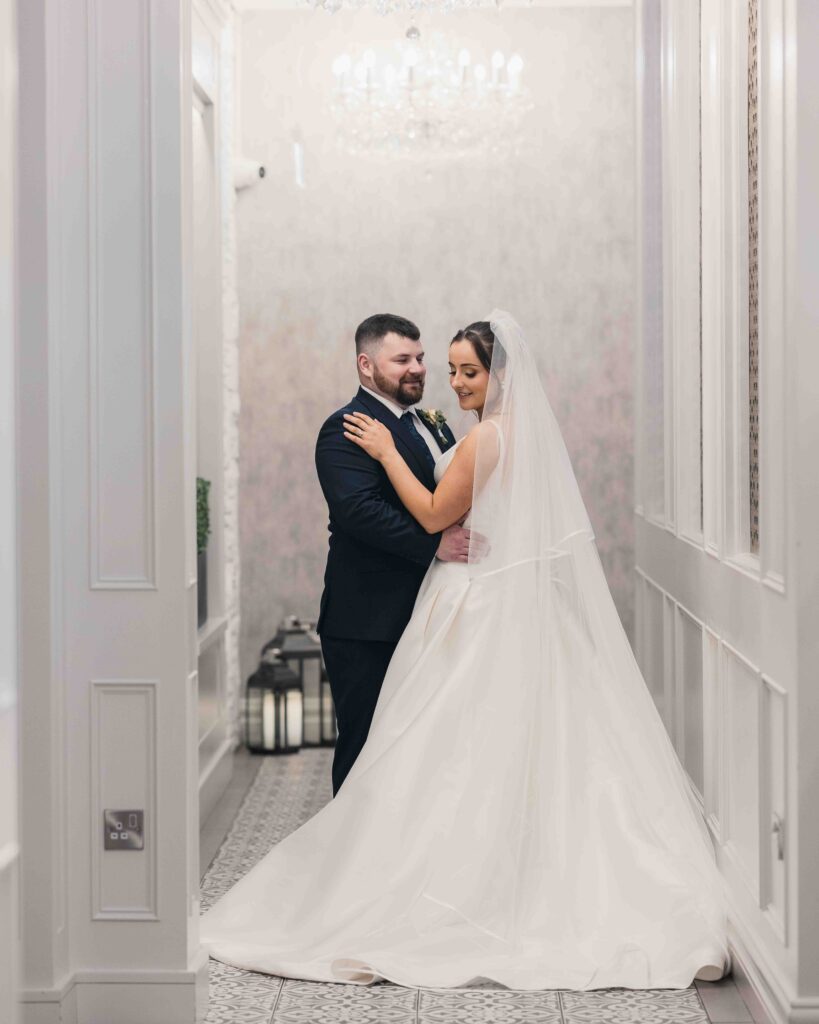 Bride and groom embracing in the white panelled hallway with a chandelier at The Oaks Hotel in Killarney