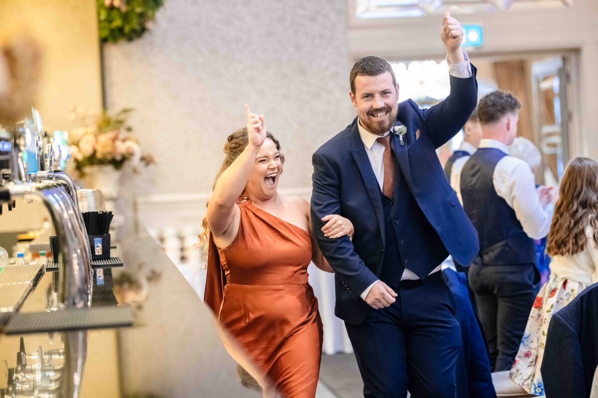 Groom in a navy suit and a bridesmaid in a rust dress cheering with arms raised as they arrive at the bar at The Oaks, Killarney