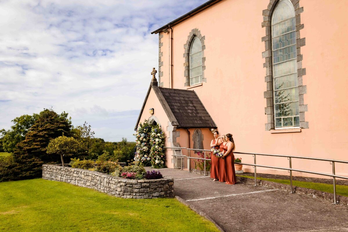 Bridesmaids in rust dresses waiting by the door of the pink Derrinagree Church before the ceremony, Co. Cork
