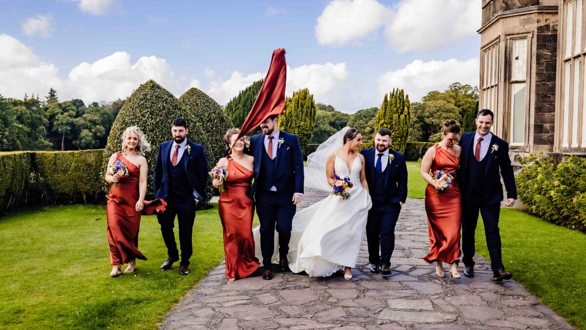 Bridal party in rust dresses and navy suits walking laughing along a paved path at Muckross House with the bride's veil blowing behind