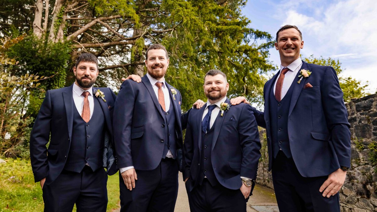 Four groomsmen in navy three-piece suits with arms around each other laughing outdoors on the wedding day