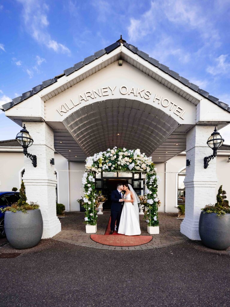Bride and groom under a white flower arch at the entrance to The Oaks Hotel in Killarney, Co. Kerry, on their wedding day