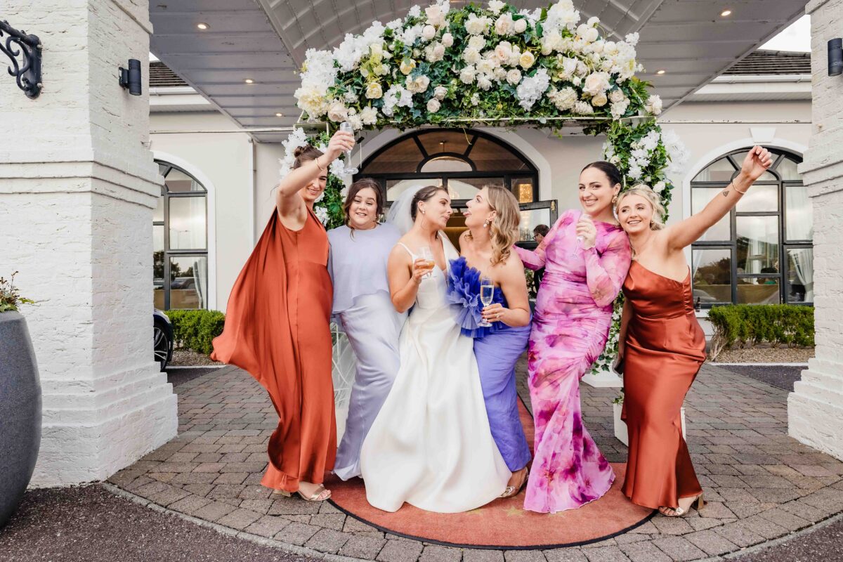 Bride and bridesmaids celebrating with their arms in the air under a flower arch at The Oaks Hotel Killarney wedding