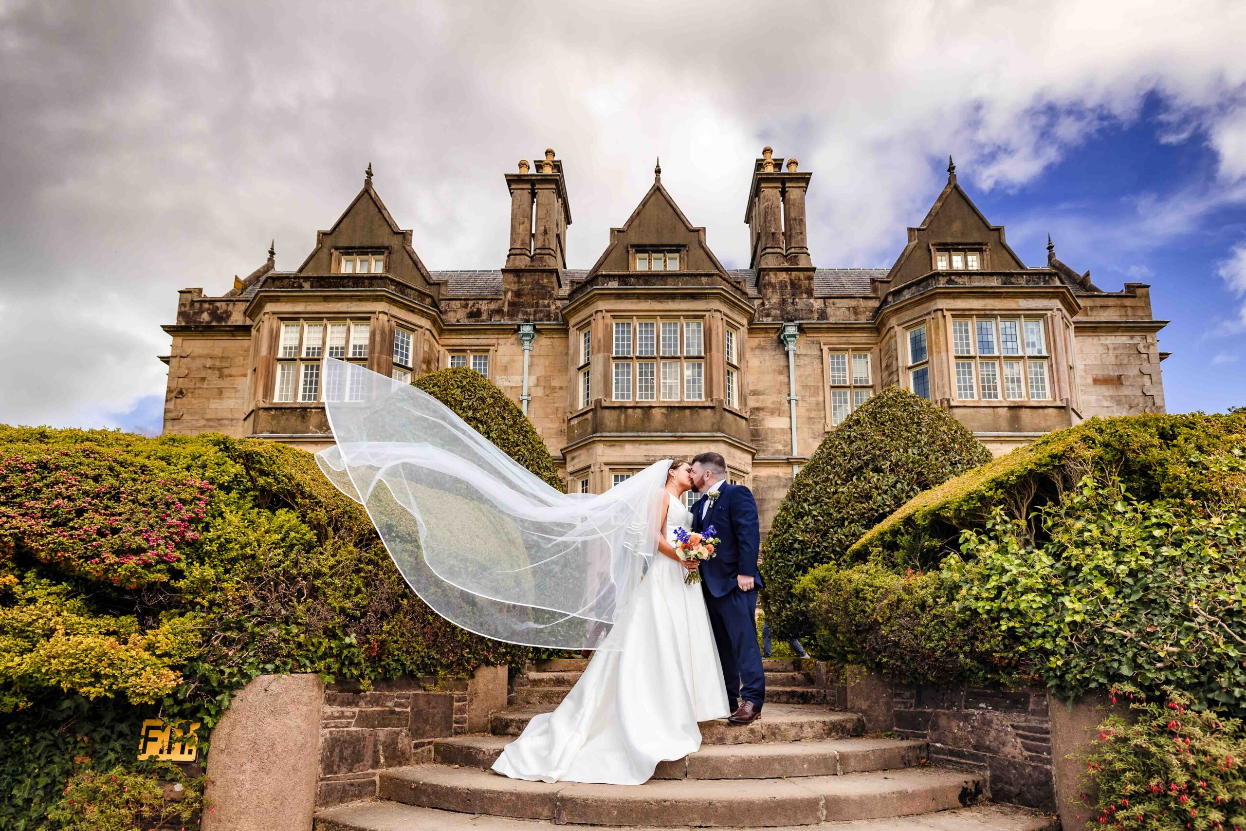 Killarney wedding photography at Muckross House, Co. Kerry: the bride's long veil blowing as she kisses the groom on the stone garden steps