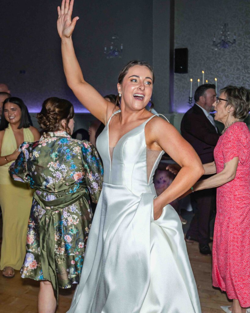 Bride in a satin wedding dress with one arm raised high dancing on the dance floor beside guests in floral and pink dresses