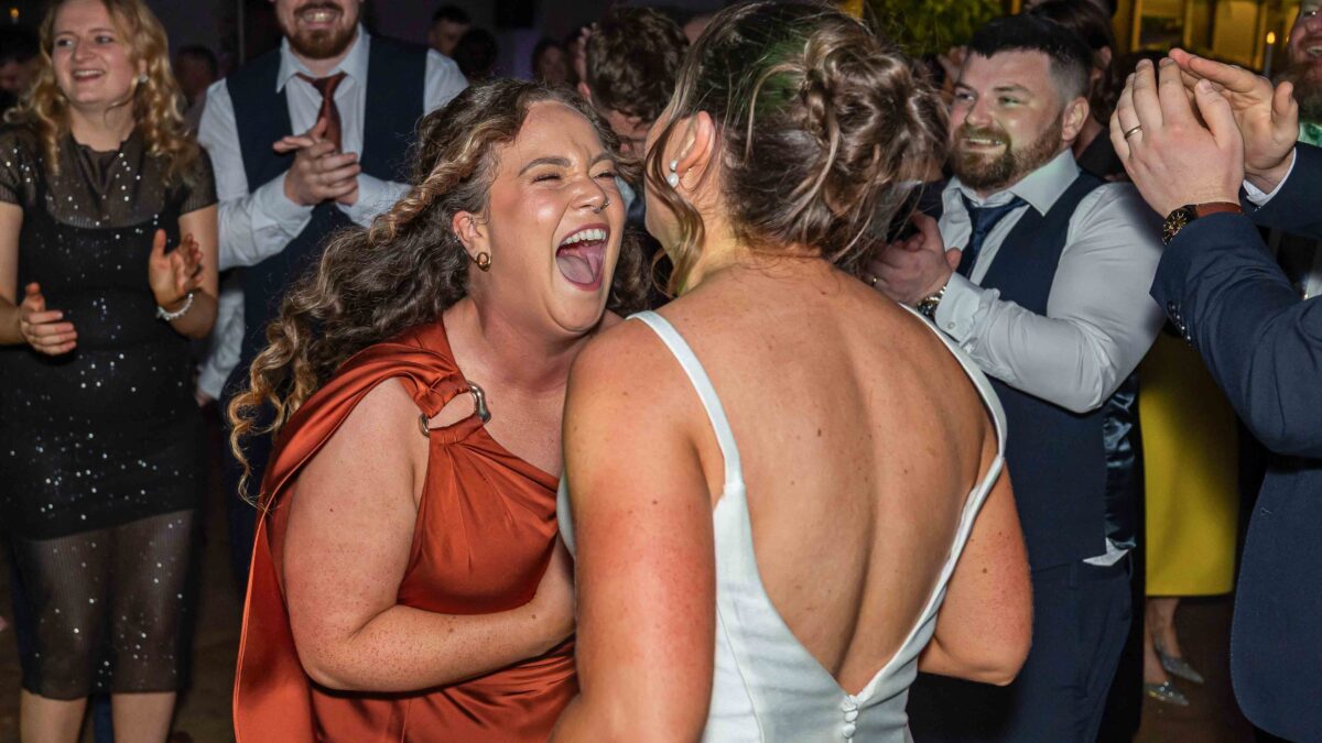 Bride in a low-back wedding dress and a bridesmaid in rust laughing together in the middle of the dance floor