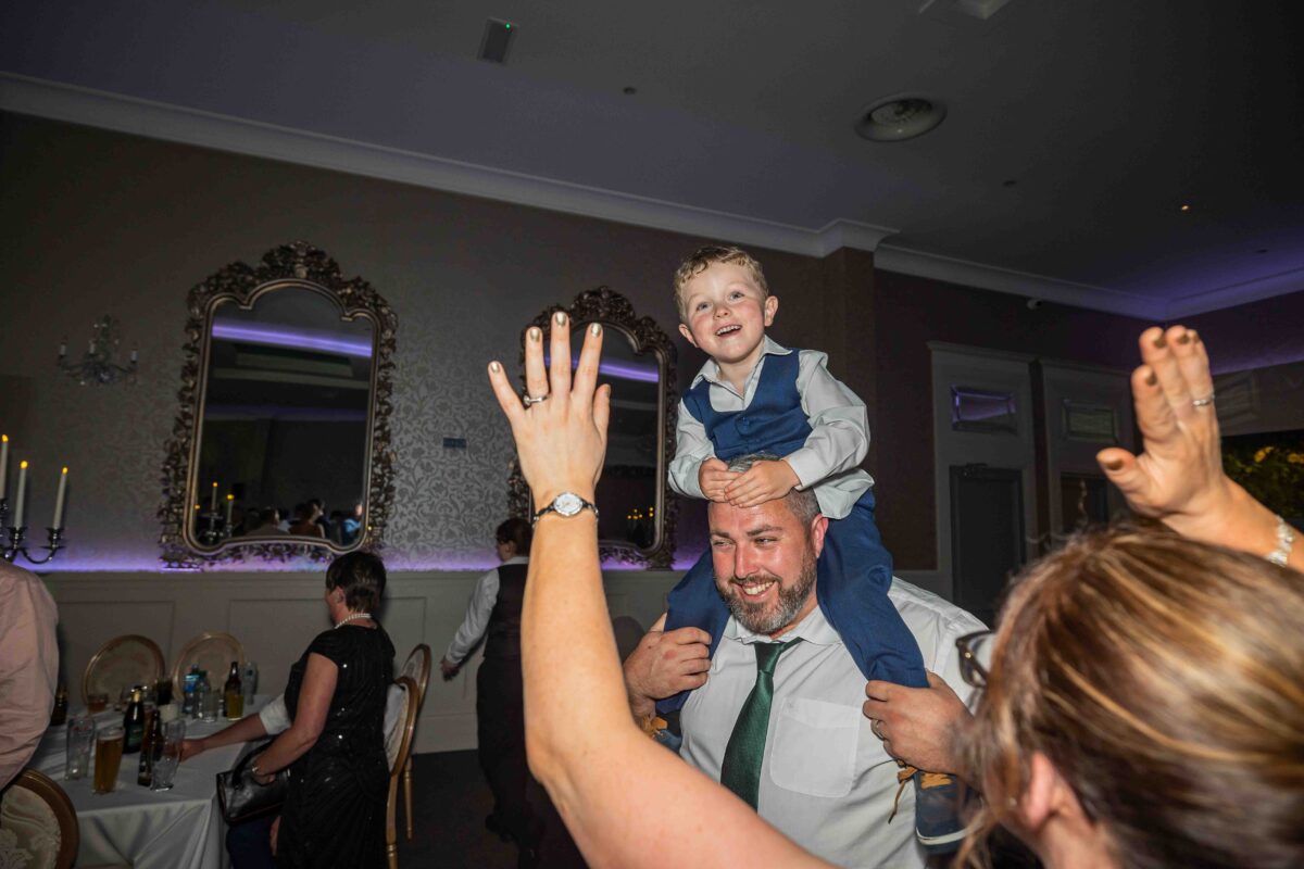 Little boy in a waistcoat on his dad's shoulders as guests dance with their arms raised in the ballroom at The Oaks, Killarney