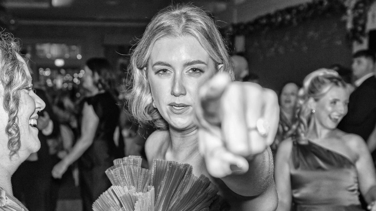 Black and white photo of a wedding guest pointing straight at the camera while holding a fan of cash on the dance floor