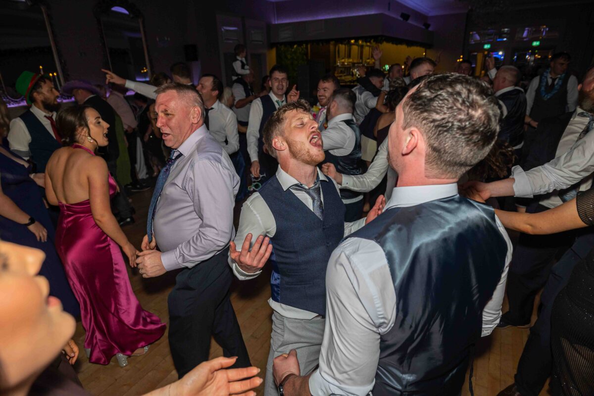 Groomsmen and guests dancing and singing in a packed crowd on the dance floor at the wedding reception