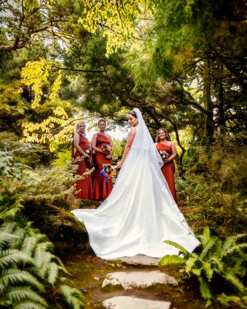 Bride in a long white gown with a flowing train on a fern-lined path in the gardens at Muckross House with three bridesmaids in rust dresses behind her