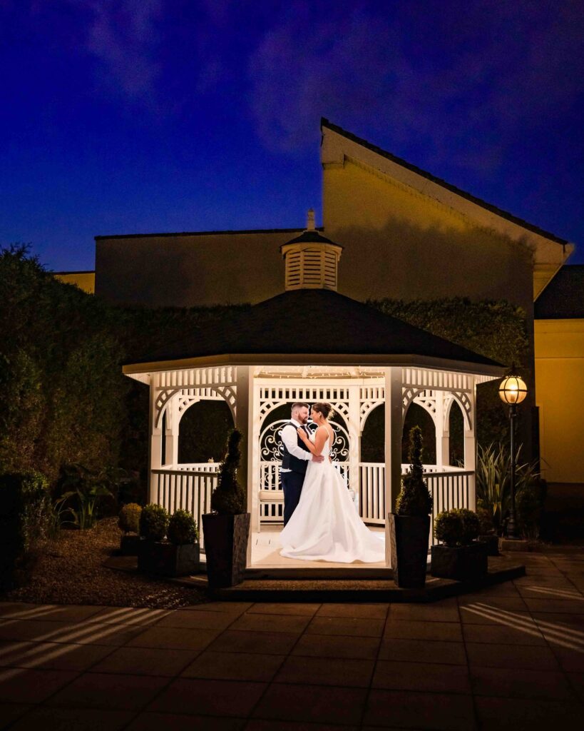 The Oaks Hotel Killarney wedding photos: bride and groom embracing inside the lit white gazebo against a deep blue night sky