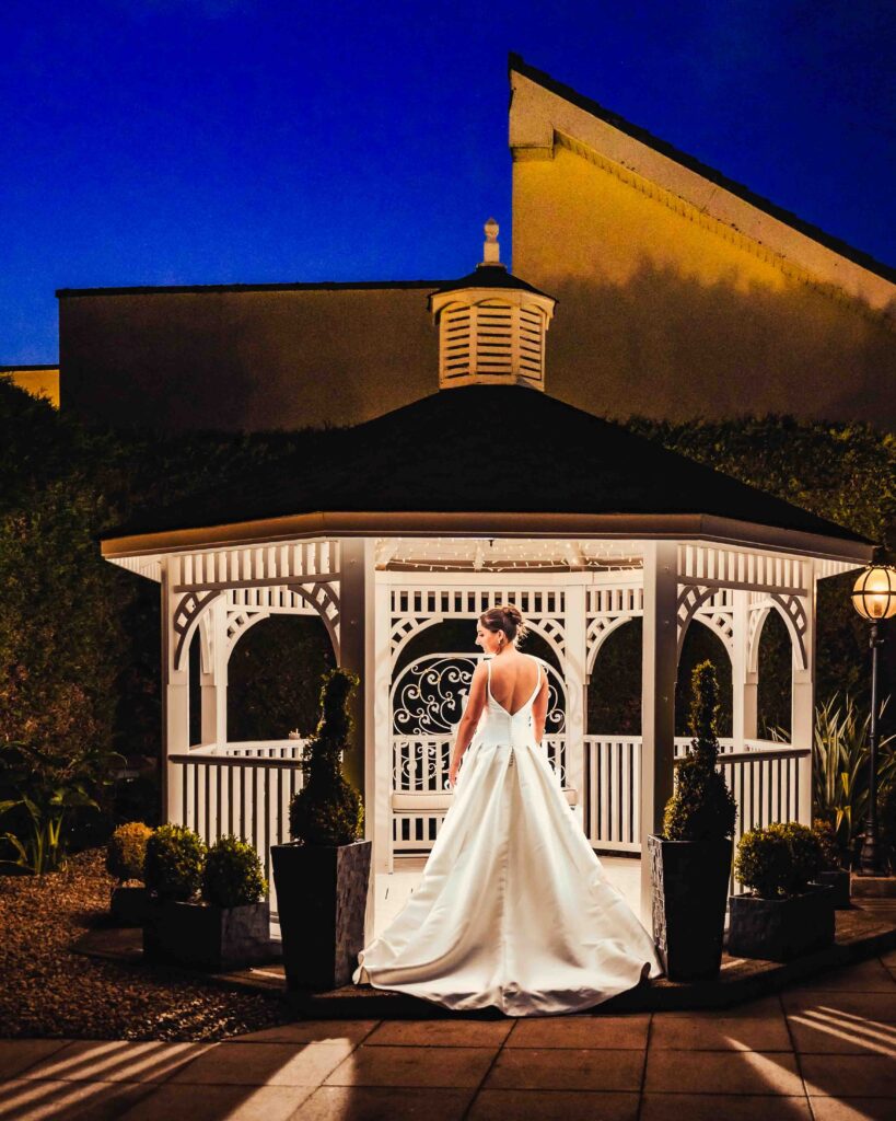 Bride in a satin wedding dress standing in the glowing white gazebo at The Oaks Hotel in Killarney at dusk
