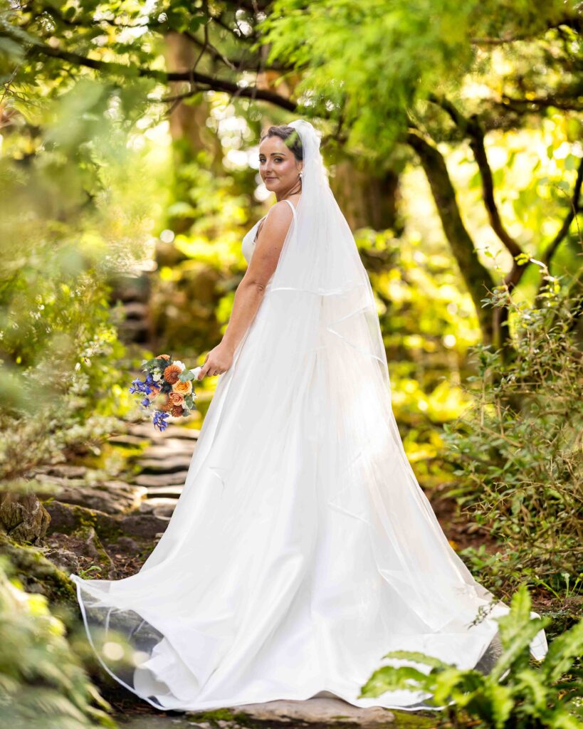 Bride looking back over her shoulder with a bouquet as her veil and dress flow on a woodland path in the gardens at Muckross House, Killarney