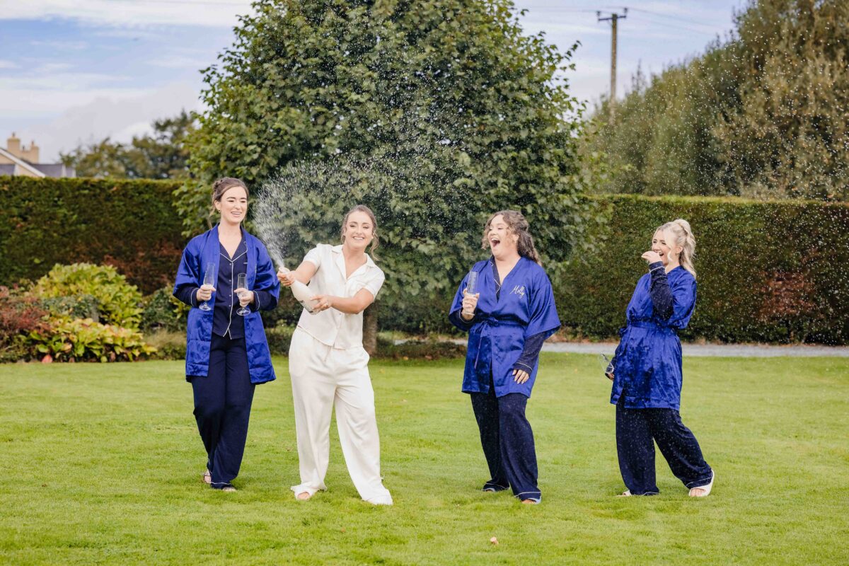 Bride in white pyjamas and three bridesmaids in navy satin robes laughing and dancing on a lawn on the wedding morning