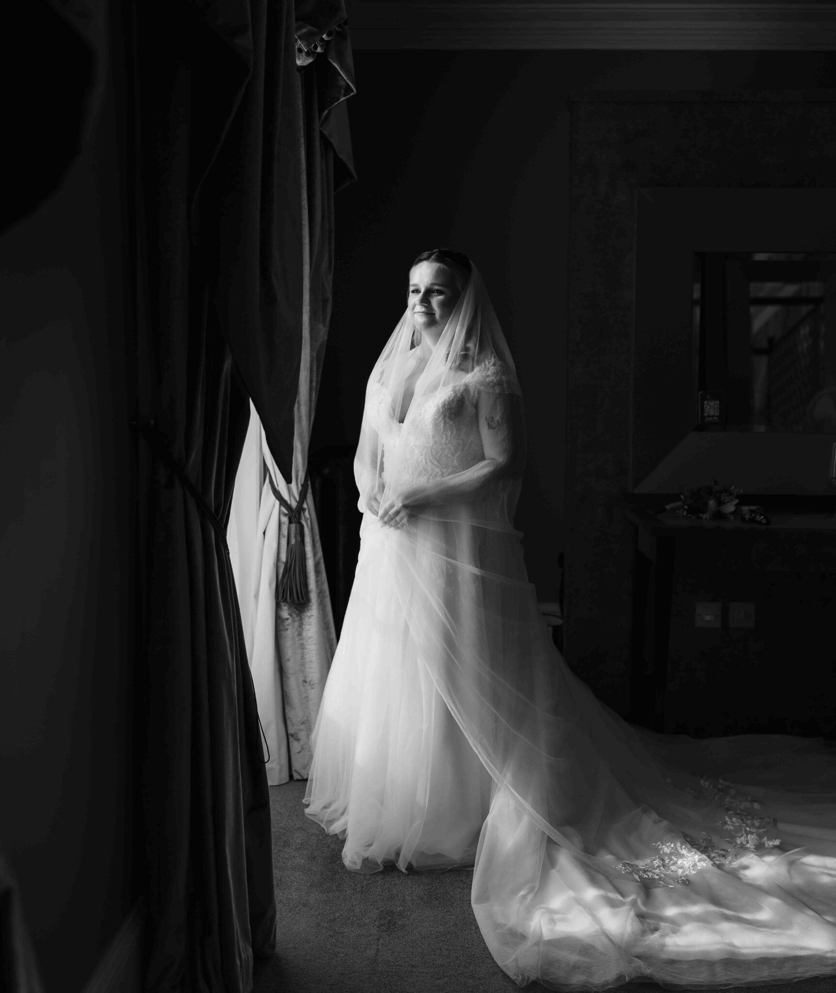 Black and white photo of Shannon in her wedding dress and veil standing in a dark window at the Talbot Hotel Cork