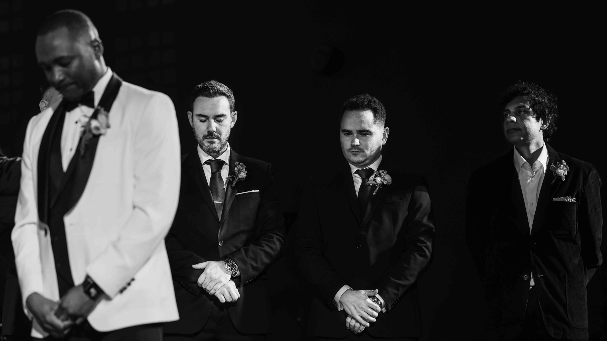 Mahan and his groomsmen stand heads bowed during the ceremony, black and white wedding photography Cork