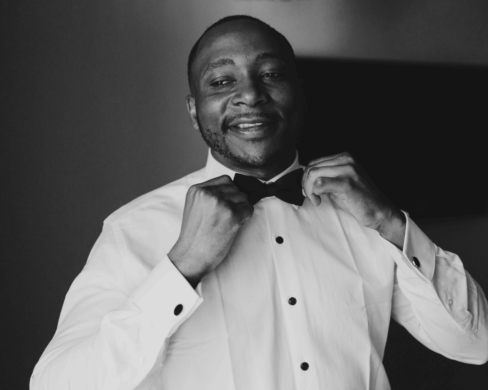 Black and white portrait of groom Mahan adjusting his bow tie and smiling before the wedding, Cork